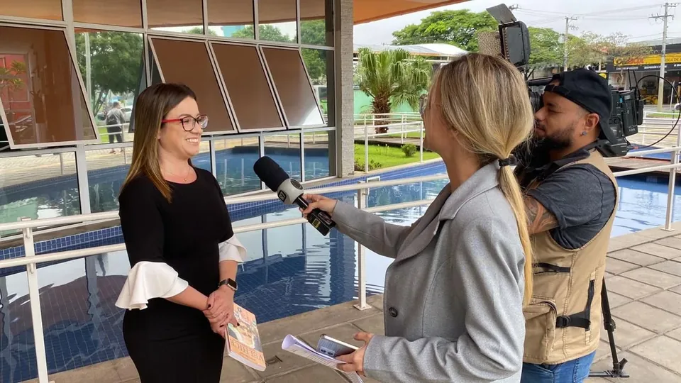 Vanessa Fortes falando durante entrevista em área externa, com microfone e câmera ao lado